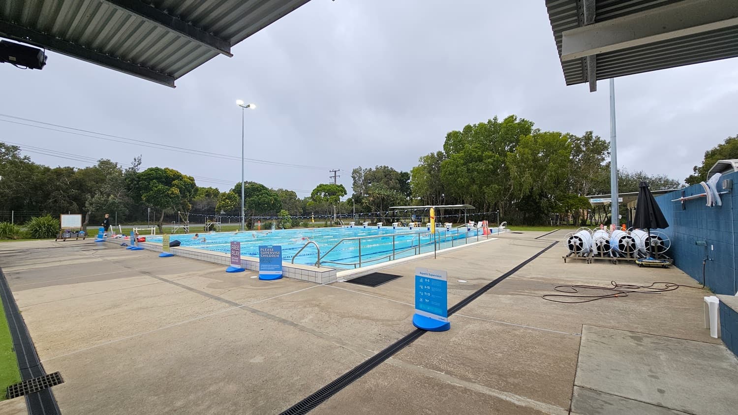 An outdoor swimming pool with clear blue water is surrounded by a concrete deck. On the right, there are several large white reels and a black umbrella against a blue wall. Signs with instructions are placed around the pool. In the background, there are tall trees and a fence. The ceiling is a metal structure extending from the top left corner. The area is well-lit with tall lights.