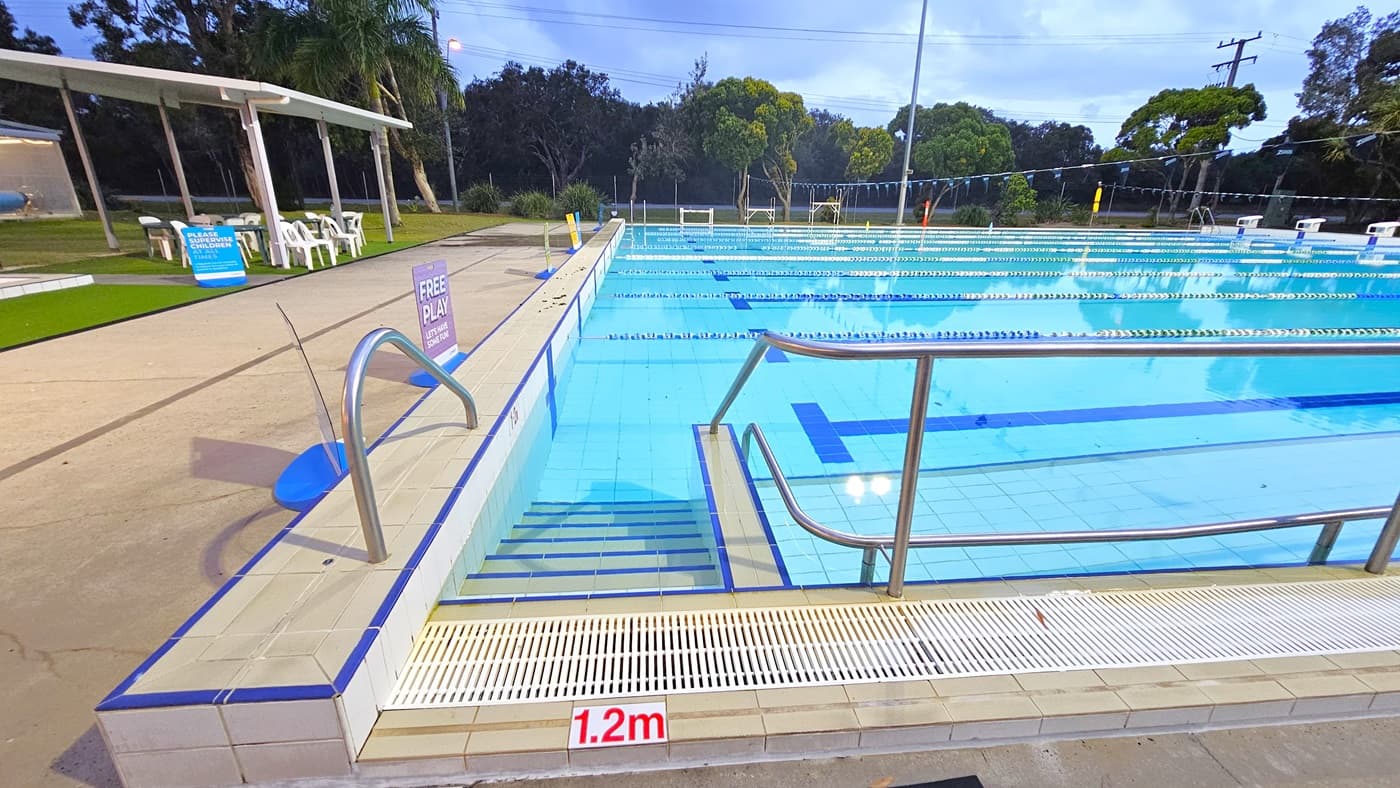 Outdoor swimming pool with clear blue water and lane dividers. On the left, there are steps with a metal handrail leading into the pool. A sign indicates a depth of 1.2 metres. In the background, there is a grassy area with white plastic chairs under a shelter. Trees and a fence line the perimeter. The pool deck is tiled, and there are signs for "Free Play" and safety notices.