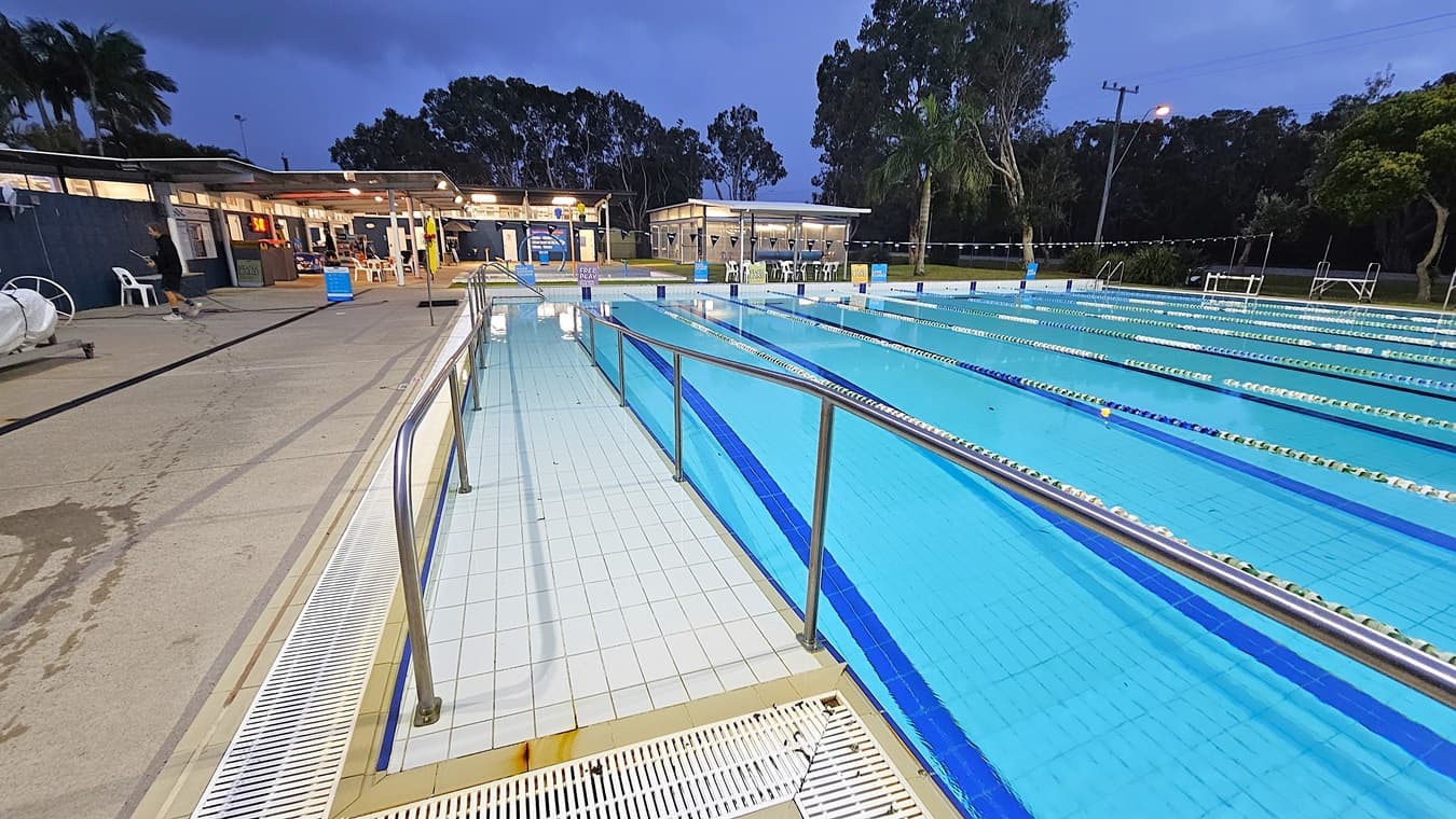 An outdoor swimming pool with multiple lanes, bordered by metal railings. The pool is on the right, with clear blue water and lane dividers. On the left, a wet concrete poolside area with chairs and a few people. In the background, there are buildings with lights on, surrounded by trees. The sky is overcast, and a streetlamp is visible near the trees.