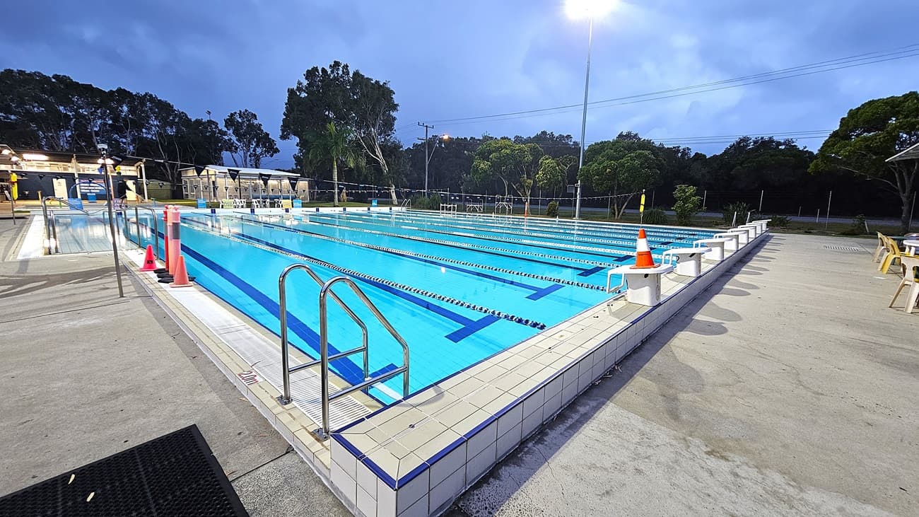 An outdoor swimming pool with clear blue water, divided into lanes by blue and white lane markers. On the left, there are metal handrails and a few orange cones on the pool deck. The right side shows more of the pool deck with some chairs and tables. In the background, there are trees and a lit area with more seating. The pool area is illuminated by tall lights, and the sky appears overcast.
