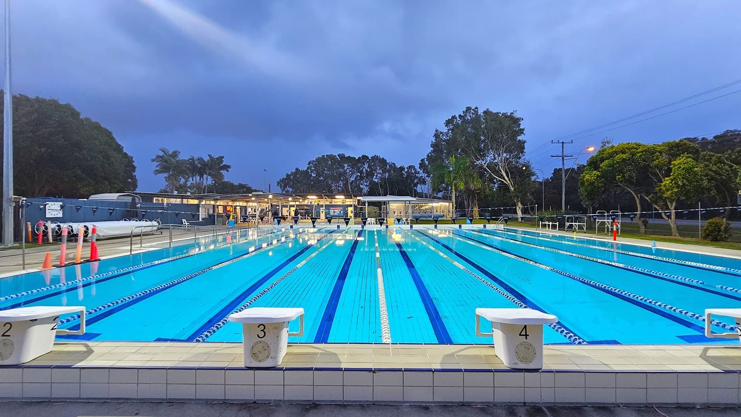 An outdoor swimming pool with multiple lanes, each marked by blue and white lane dividers. Starting blocks numbered 2, 3, and 4 are visible in the foreground. The pool is surrounded by a tiled deck. In the background, there are trees and a building with lights on, possibly a changing facility. A few orange cones are placed near the poolside. The scene is set during twilight, with a darkening sky.