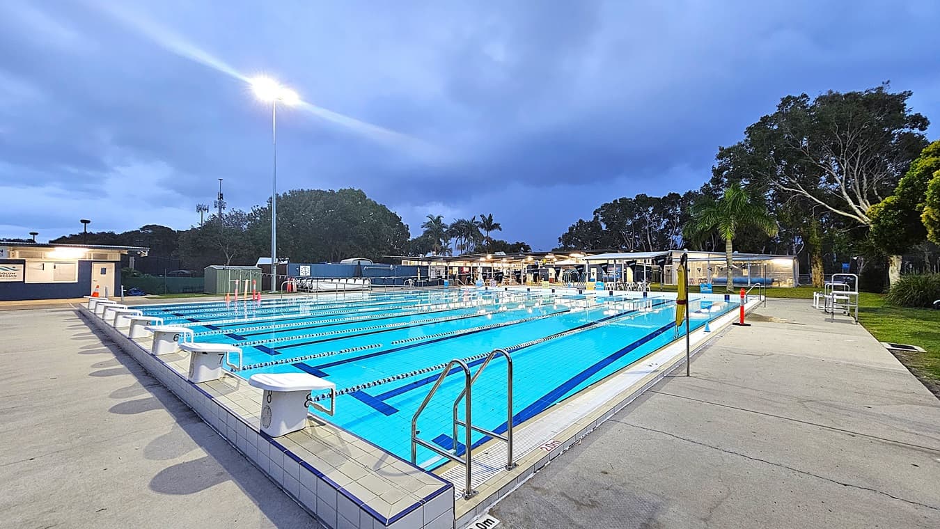 An outdoor swimming pool with multiple lanes, surrounded by a tiled deck. Starting blocks are positioned at the pool's edge. On the left, a building with a lit entrance is visible. In the background, there are trees and a few palm trees, with a tall light pole illuminating the area. The pool area is bordered by a concrete path, and there are some chairs and equipment near the poolside.