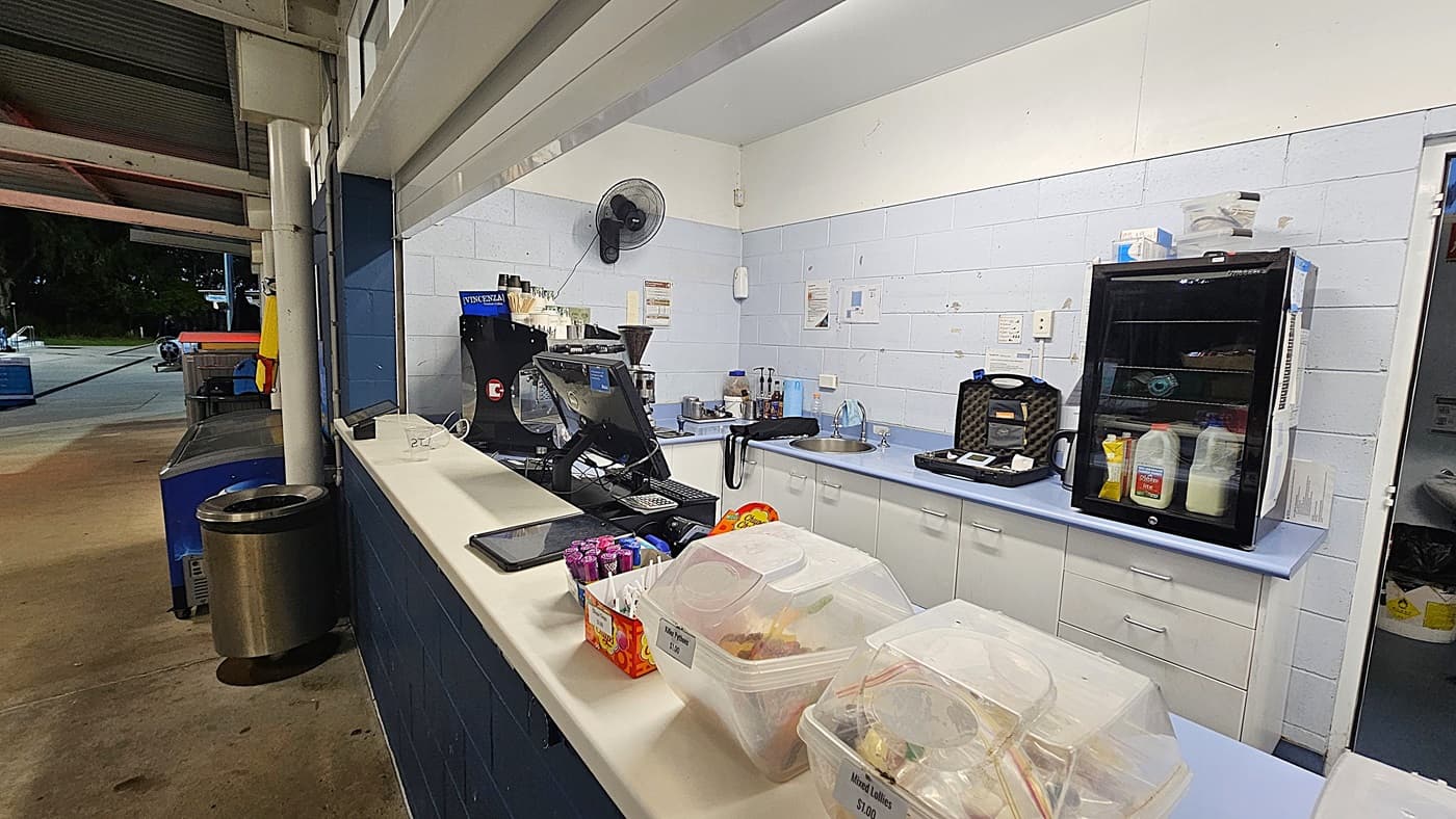 A small café counter with a coffee machine on the left, a computer monitor in the centre, and various snacks and drinks displayed on the counter. Behind the counter, there are shelves with condiments and a refrigerator. To the right, a vending machine and a display stand with snacks are visible. A sign on a pillar reads, "DIRECT PARENT SUPERVISION IS THE ONLY SUPERVISION." The walls are light blue, and the counter is dark blue.