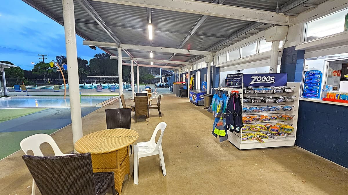 A covered outdoor area with a concrete floor. On the right, there's a display rack with swimming goggles and accessories labelled "Zoggs". Nearby, a counter with more items and a menu board. In the middle, tables and chairs are arranged for seating. On the left, a swimming pool is visible with water play features and trees in the background. The ceiling is metal with fluorescent lights.