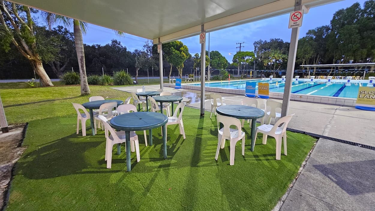 Under a covered area, there are several green tables with white plastic chairs on artificial grass. To the right, a swimming pool with marked lanes is visible, surrounded by a concrete path. Signs indicate "Lane Closed" and "Slow Lane". In the background, there are trees and a grassy area with a few palm trees. The ceiling is a simple flat structure supported by metal poles.