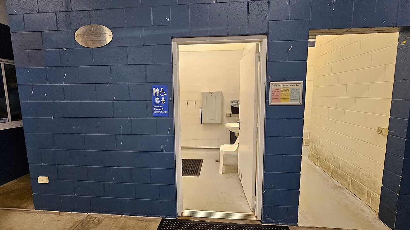 A public restroom entrance with a blue brick wall. On the left, a plaque is mounted above a sign indicating facilities for toilets, showers, and baby changing. The open door reveals a white interior with a sink, a chair, and a baby changing station. To the right, a sign displays "Parent Supervision Rules" next to a hallway with light-coloured brick walls. The floor is concrete, and a black mat is placed at the entrance.