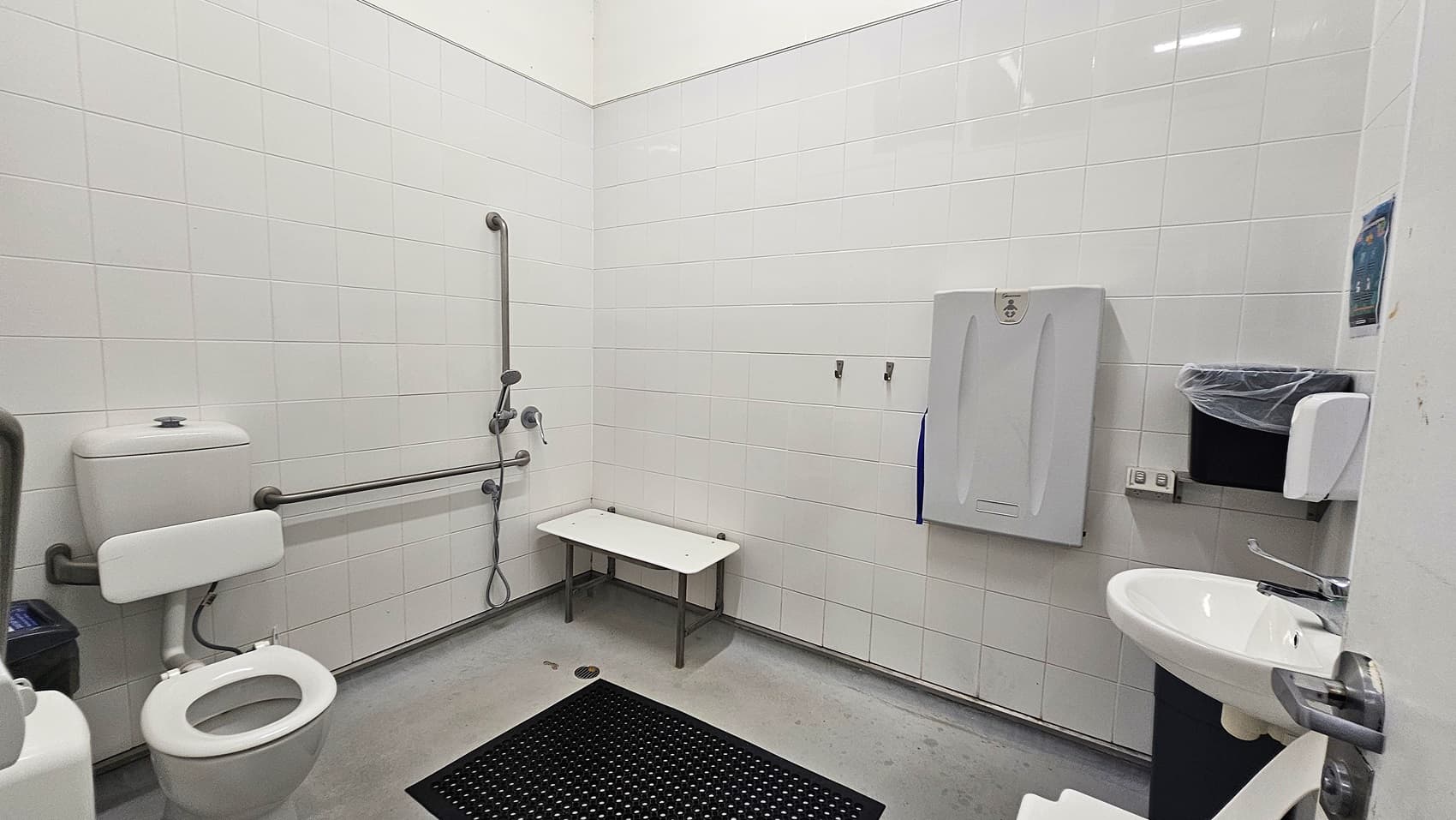 A spacious, accessible bathroom with white tiled walls and a grey floor. On the left, there's a toilet with support rails. In the centre, a shower area with a foldable bench and additional rails. On the right, a baby changing station is mounted on the wall above a bin. A sink with a lever tap is near the door, which is partially open. A black non-slip mat is on the floor.