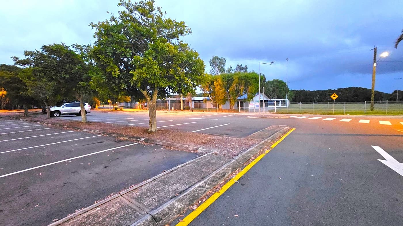 A dimly lit street scene at dusk with a zebra crossing in the foreground. On the right, there's a building with illuminated windows and several tall trees. A streetlamp casts a warm glow over the pavement. On the left, a fence with banners lines the road, and trees are visible in the background. The road curves slightly, leading into the distance under a deep blue sky.