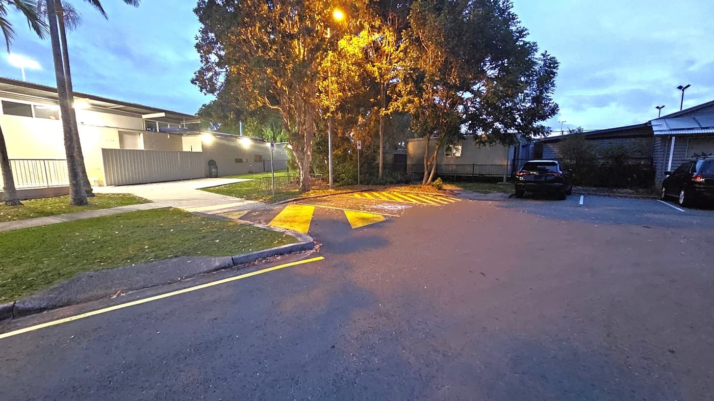 A parking area with two cars on the right. In the centre, a tree is illuminated by a streetlamp, casting a warm glow. Yellow directional arrows are painted on the ground. On the left, a building with a flat roof and lit windows is visible, with a small grassy area and a footpath leading to it. The sky is dusky, suggesting early evening.