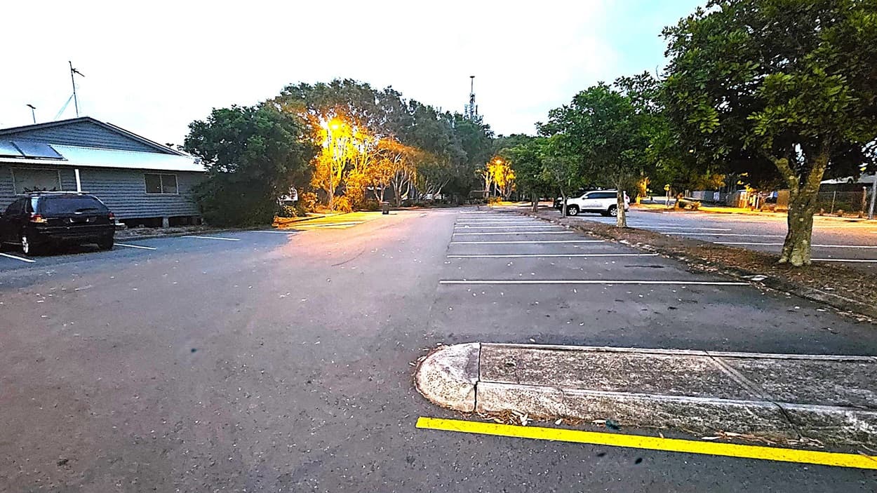 A nearly empty car park with marked spaces. On the left, a dark-coloured car is parked next to a building with a sloped roof and solar panels. Trees line the background, illuminated by streetlights casting a warm glow. On the right, a white car is parked near more trees. The ground is asphalt with some scattered leaves, and a concrete kerb with a yellow line is visible in the foreground.