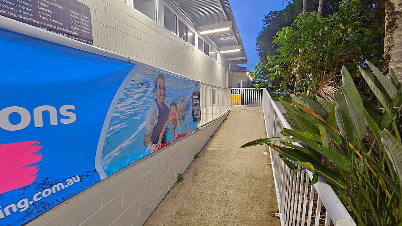 A pathway with a light-coloured concrete surface runs between a building on the left and a row of plants on the right. The building has a light green brick wall with a large banner showing a swimming lesson. Above, there are windows and a metal roof with lights. A white railing lines the path, and there are trees in the background.