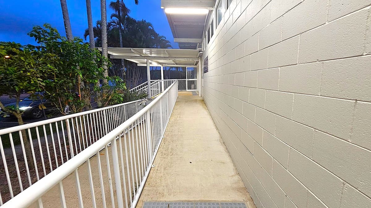 A concrete ramp with white metal railings on both sides leads to a building entrance. On the right, there's a light green brick wall. On the left, there are trees and a parked car partially visible. The ceiling above the ramp is covered with a transparent roof, and fluorescent lights illuminate the area.