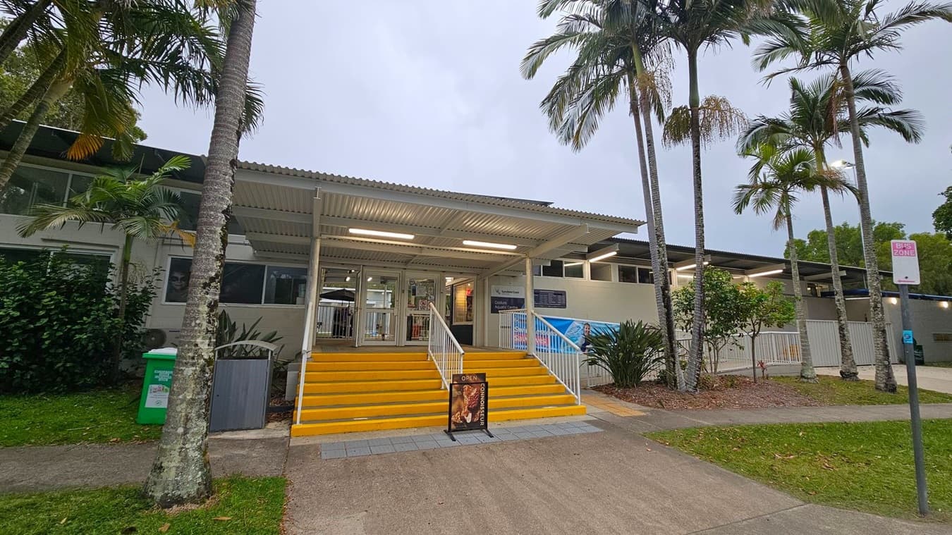 Entrance to a building with a corrugated metal roof and yellow steps leading up to glass doors. A signboard at the bottom of the steps reads "OPEN." Palm trees and greenery are on both sides, with a green bin on the left. A "BUS ZONE" sign is on the right. The building has large windows and a light green exterior. The ground is a mix of concrete and grass.