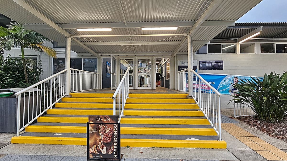 A building entrance with a set of bright yellow steps leading up to glass doors. The steps are flanked by white railings. On the left, there's a small palm tree and a green bin. On the right, a large plant and a banner featuring a person in water. Above, a corrugated metal roof with fluorescent lights. A sign at the bottom of the steps advertises ice cream.