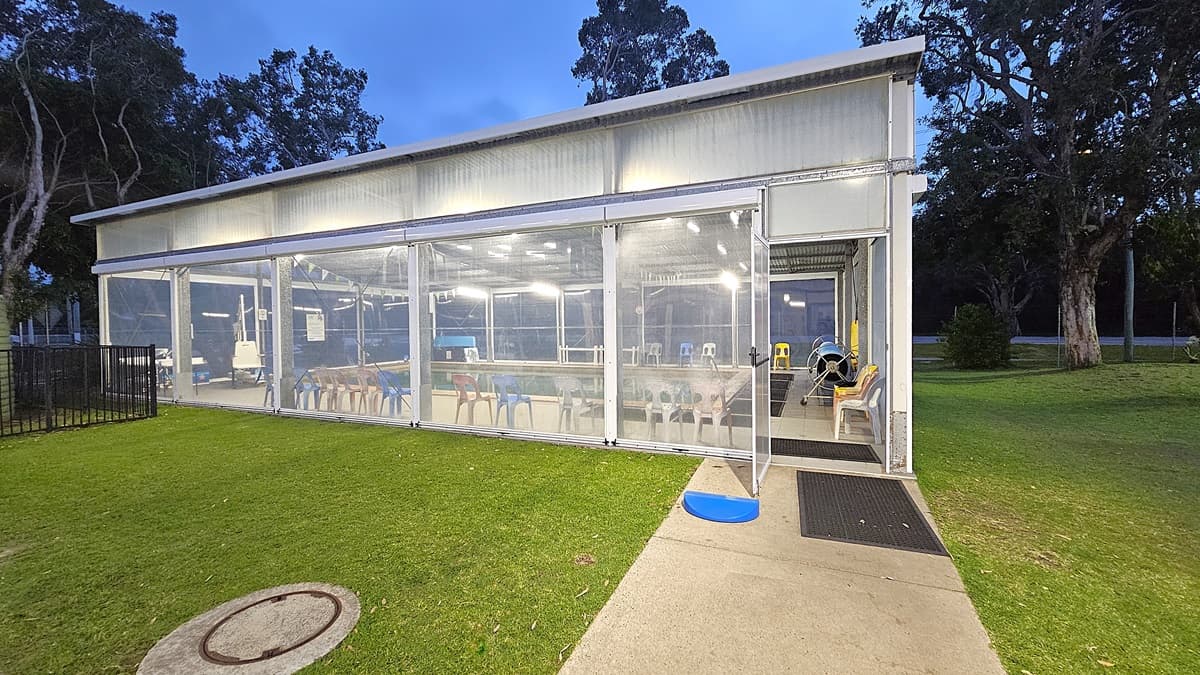 A transparent, enclosed structure with a flat roof is situated on a grassy area. Inside, there are tables and colourful chairs visible through the clear walls. The entrance on the right has a small blue step and a black mat. Trees surround the building, and the ground is a mix of grass and a concrete path leading to the entrance. The interior is well-lit, and the ceiling has visible lighting fixtures.
