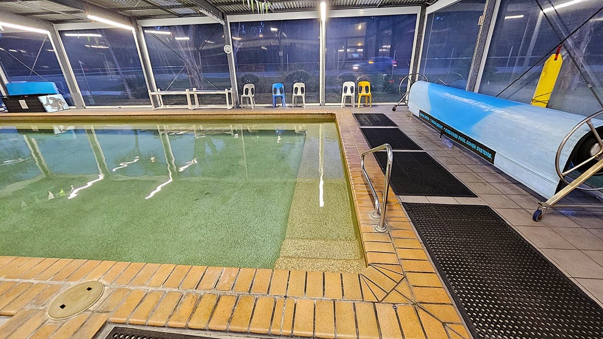 Indoor swimming pool with clear water, surrounded by a brick edge. On the right, a metal handrail leads into the pool. A blue pool cover is rolled up on a metal frame beside the pool. Several plastic chairs in different colours (blue, white, yellow) are lined up against the glass wall in the background. The floor is tiled with black rubber mats placed along the edge. The ceiling is made of corrugated metal with fluorescent lights.