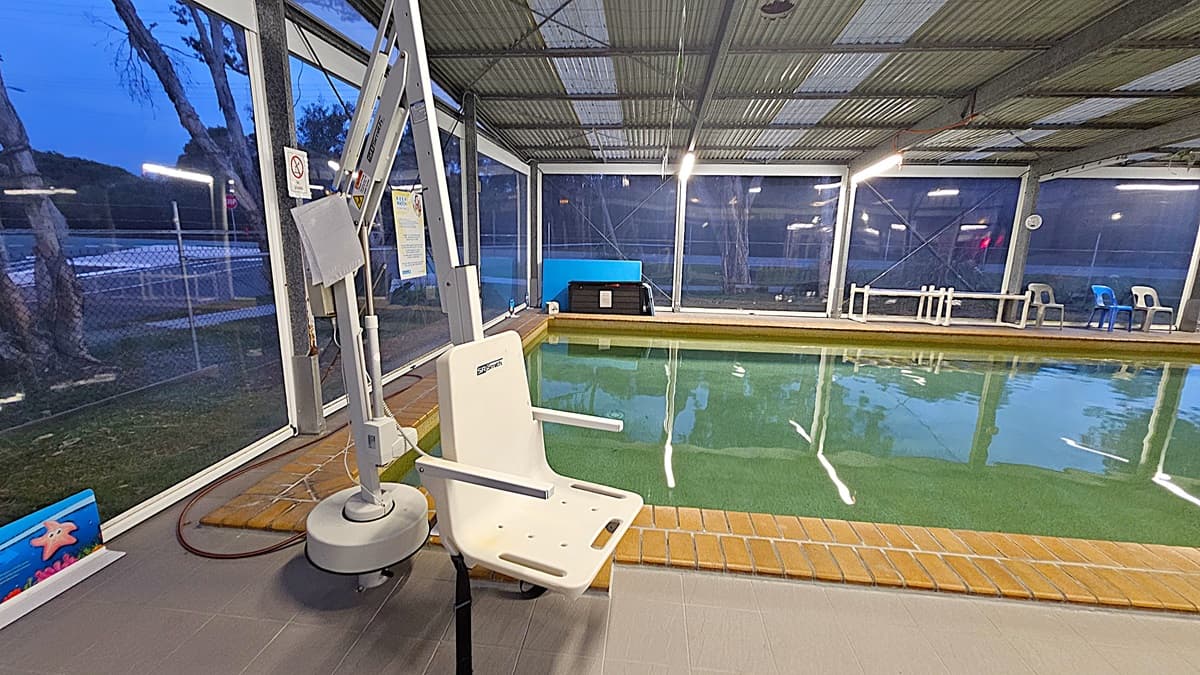 An indoor swimming pool area with a mechanical pool lift chair on the left, positioned on a tiled floor. The pool has a brick edge and clear water. Large windows line the background, showing a view of trees and a fence outside. On the right, there are a few plastic chairs in blue and grey. The ceiling is made of corrugated metal with fluorescent lights.