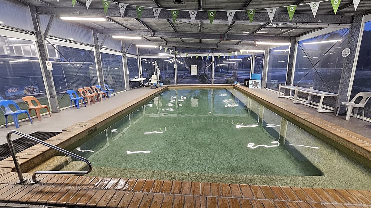 An indoor swimming pool with clear water, surrounded by a brick edge. On the left, there are orange and blue plastic chairs lined up against a wall with large windows. On the right, white plastic chairs and a long white table are visible. The ceiling is metal with fluorescent lights and green and white triangular flags hanging across. The floor is tiled around the pool area.