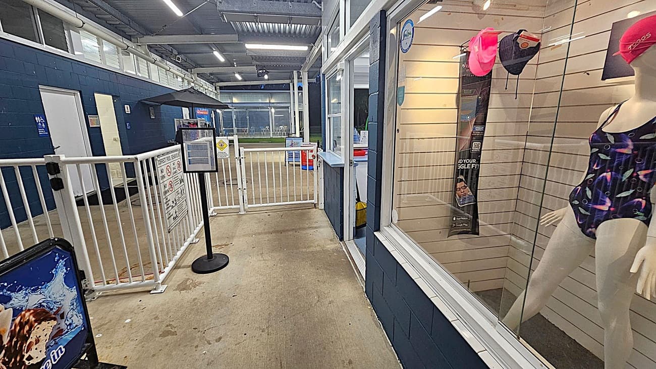 A set of glass doors with white frames, one open on the left. The doors have various signs, including "No Smoking" and "No Glassware." Behind the doors, a corridor with blue walls and a mannequin wearing a swimsuit and red cap on the right. A notice board with blue background is visible through the open door. The floor is concrete with textured mats near the entrance, and the ceiling is corrugated metal.