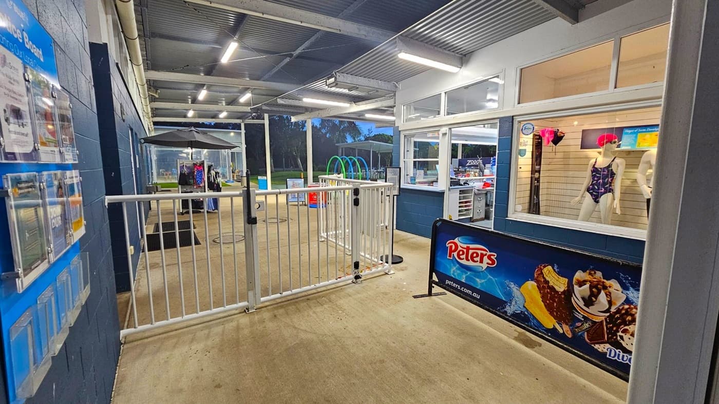 A covered outdoor area with a concrete floor and a corrugated metal ceiling. On the left, a notice board is mounted on a blue wall. In the centre, a white gate leads to a pool area with a black umbrella and clothing rack. On the right, a shop window displays mannequins in swimwear. A sign for Peters ice cream is visible below the window. In the background, there are trees and a grassy area.