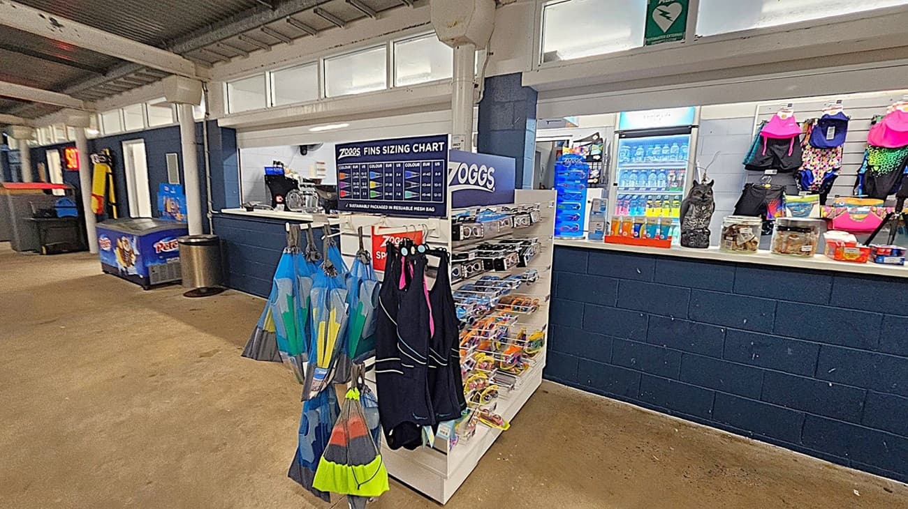 A shop interior with a display on the left featuring various swimming goggles under the brand "Zoggs". The counter on the right has snacks, a computer monitor, and a framed notice. Behind the counter, a fridge stocked with drinks is visible, along with swimwear and accessories hanging on the wall. The ceiling is metal, and the floor is concrete.
