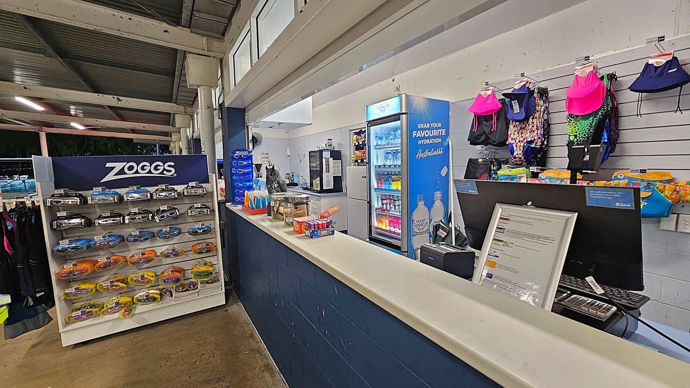 A shop area with a display stand in the centre featuring swimwear and accessories, including fins and goggles. Above the stand is a Zoggs fins sizing chart. To the right, a counter displays various items, including a jar and a decorative owl. Behind the counter, shelves hold colourful swimwear and a fridge with drinks. On the left, there's a bin and a freezer with a Peppa Pig design. The floor is concrete, and the ceiling has exposed beams.