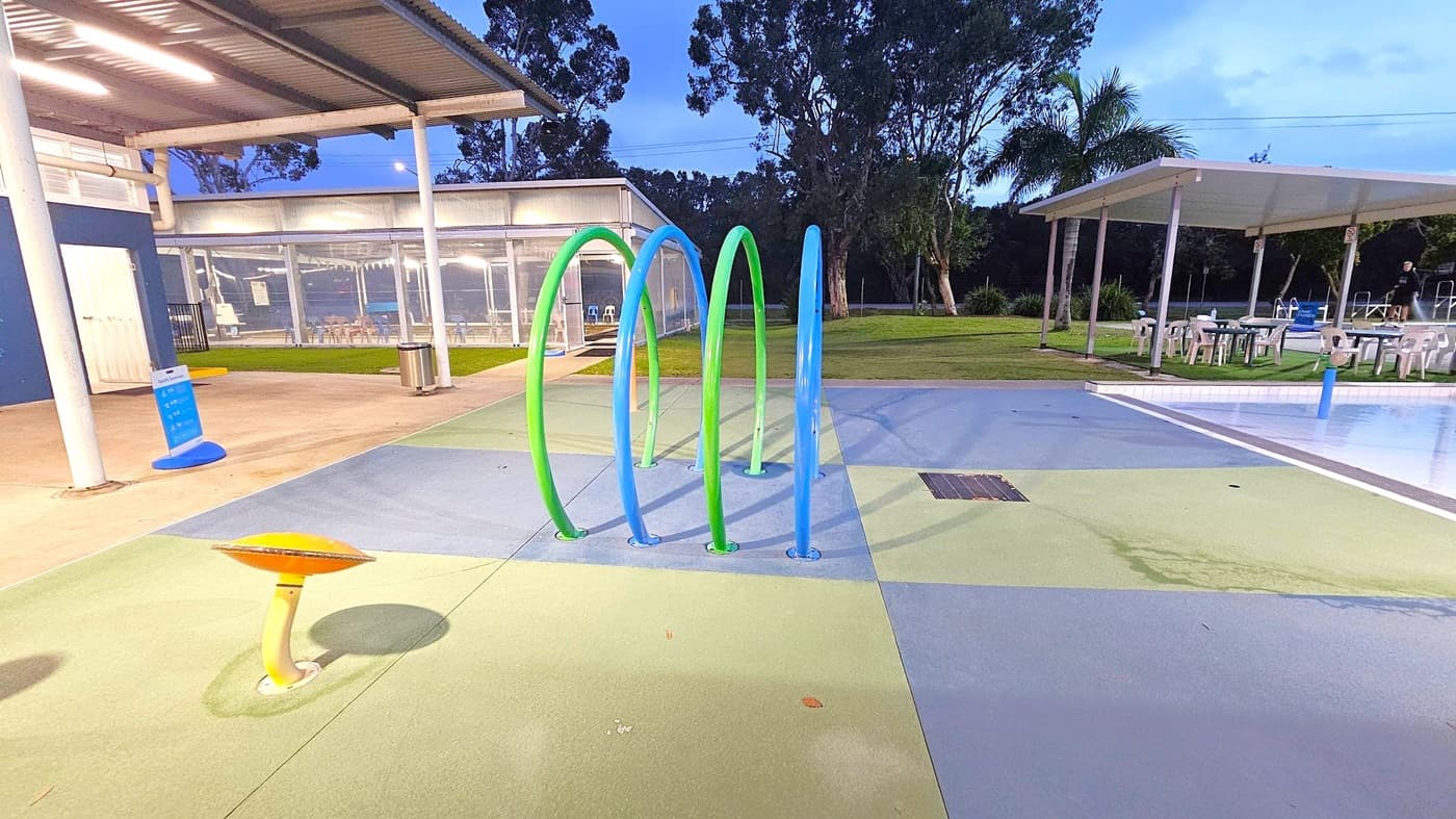 A children's water play area with colourful arches in green and blue on the left. In the foreground, a yellow post with a red top. To the right, a covered seating area with white plastic chairs and tables. In the background, a shallow pool with water features and more seating. Trees and a grassy area are visible beyond the play area. The ground is a mix of green, blue, and beige surfaces.