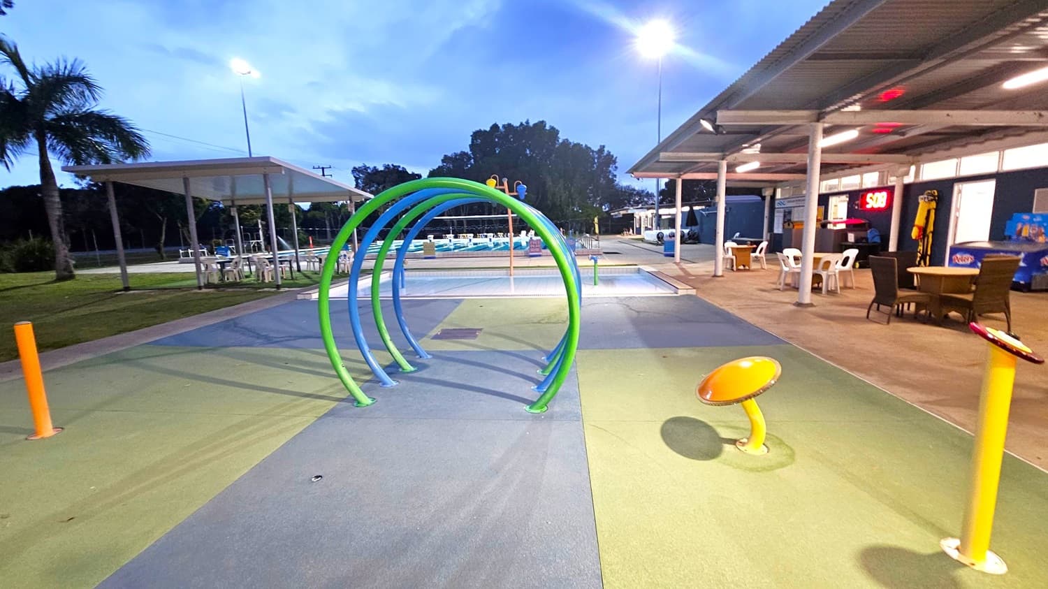 A playground area with colourful arches in the centre, set on a multicoloured rubber floor. On the right, there is a covered seating area with tables and chairs, and a digital clock displaying "5:08". On the left, a palm tree and another covered seating area with white tables and chairs are visible. In the background, there are trees and a swimming pool. The area is lit by overhead lights.