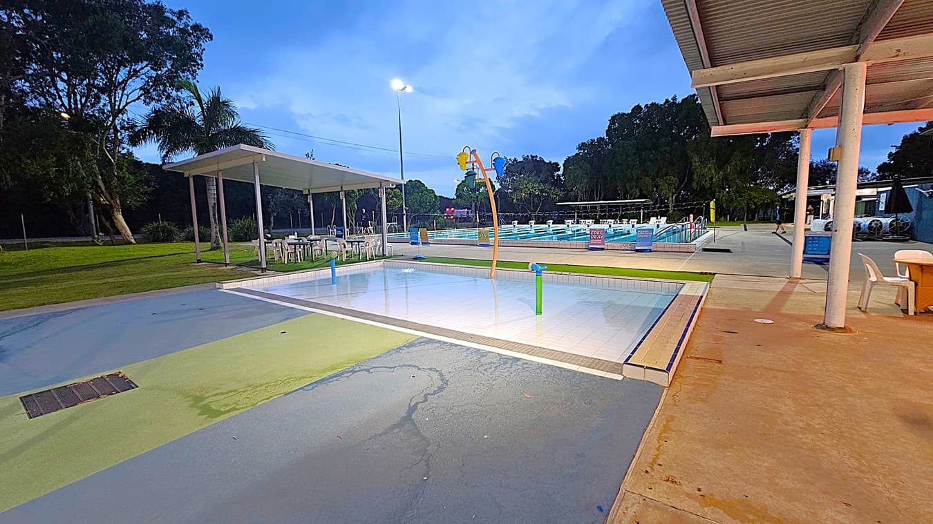 A swimming pool area with a shallow children's pool in the foreground, featuring colourful water play structures. To the left, there is a grassy area with trees and a covered seating area with tables and chairs. In the background, a larger swimming pool is visible, with lane dividers and signs. The ground is a mix of concrete and painted surfaces, and a metal-roofed structure is on the right. The scene is lit by overhead lights.