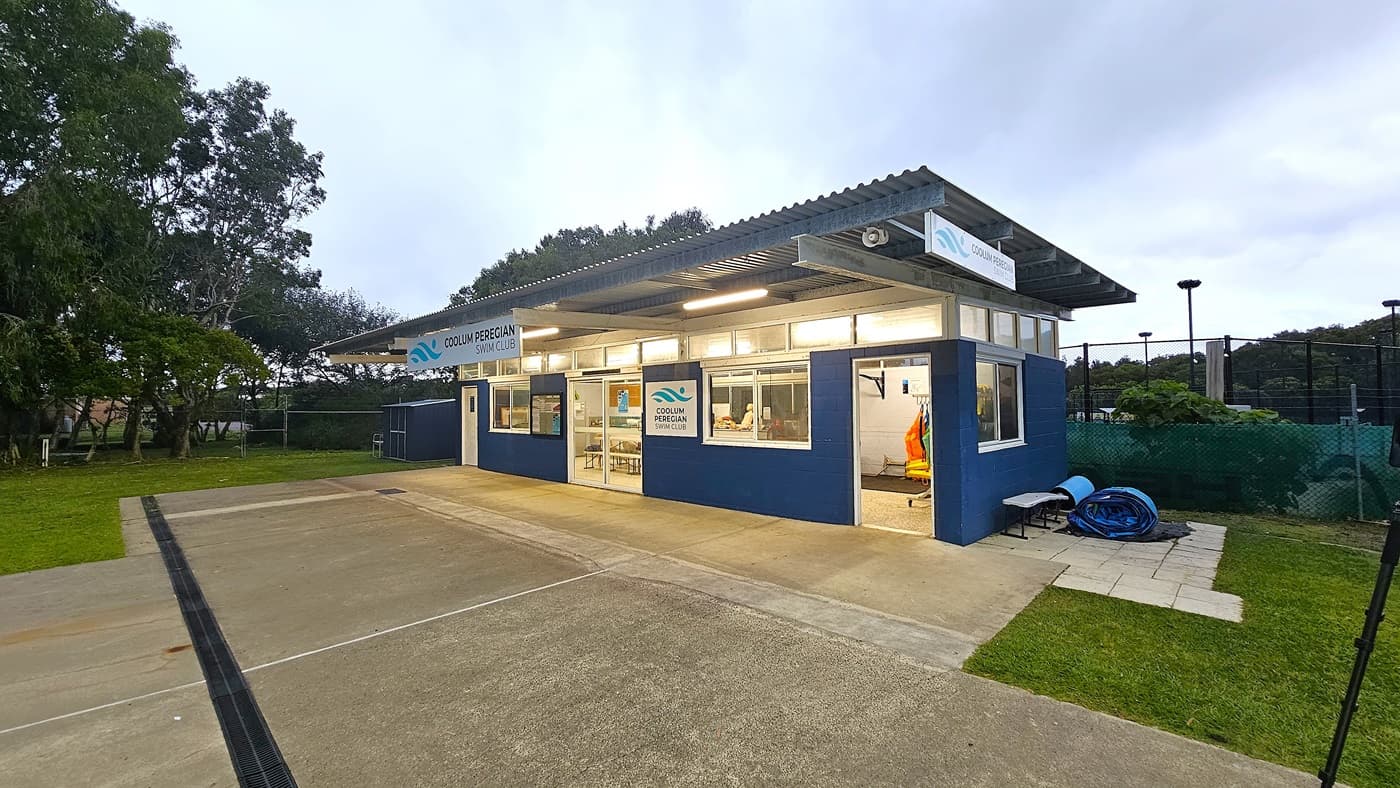 A small building with a corrugated metal roof and large windows, labelled "Coolum Peregian Swim Club". The structure is painted blue and white. Inside, there are visible tables and chairs. To the right, a rolled-up blue hose and a bench are on a paved area. The foreground has a concrete path leading to the entrance, and the background features trees and a fenced area.