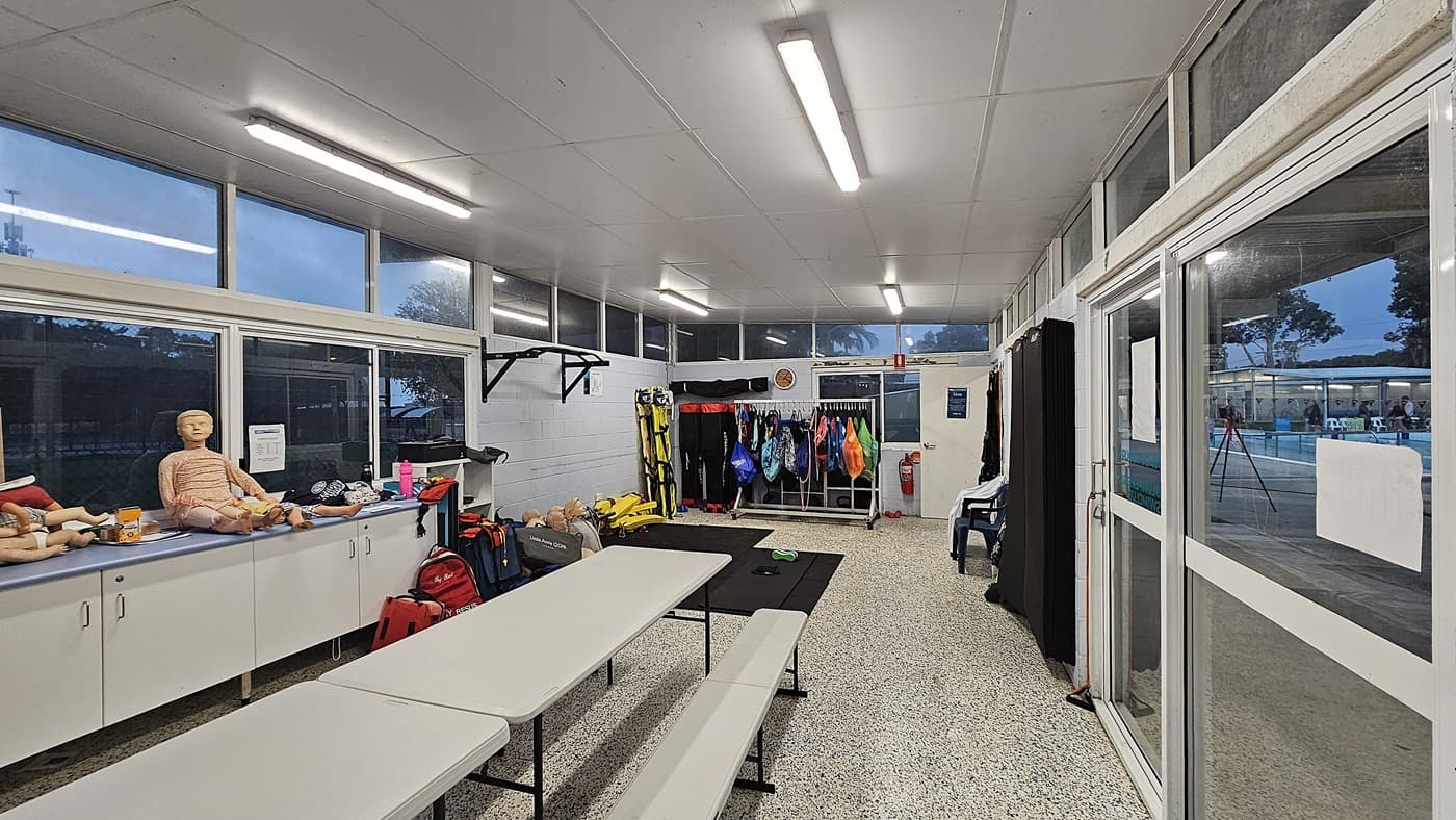 A room with a speckled floor and a white ceiling. On the left, there are cabinets with mannequins and various equipment on top. In the centre, a table with benches is placed on the floor. The right side features large windows and a glass door, revealing an outdoor pool area. The back wall has hanging racks with colourful life vests and other gear. Fluorescent lights illuminate the room.