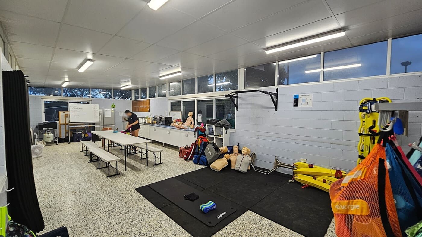 A room with a speckled floor and a white ceiling. On the left, there are long tables with benches and a person standing near them. A whiteboard and various equipment are against the wall. The right side has a wall with windows above, a pull-up bar, and several CPR mannequins on the floor. Brightly coloured bags and yellow rescue boards are hanging and stacked nearby. The room is well-lit with fluorescent lights.