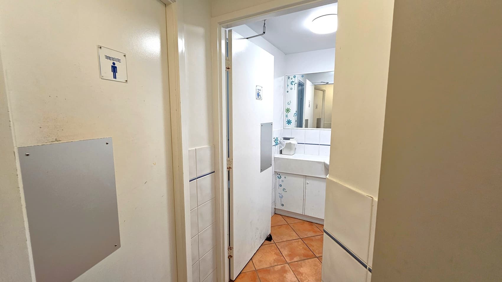 A bathroom entrance with a partially open door on the left, displaying a sign with a figure. Inside, a white sink with a paper towel dispenser is visible, set against a wall with floral tile patterns. The floor is covered with terracotta tiles, and a round ceiling light illuminates the space.