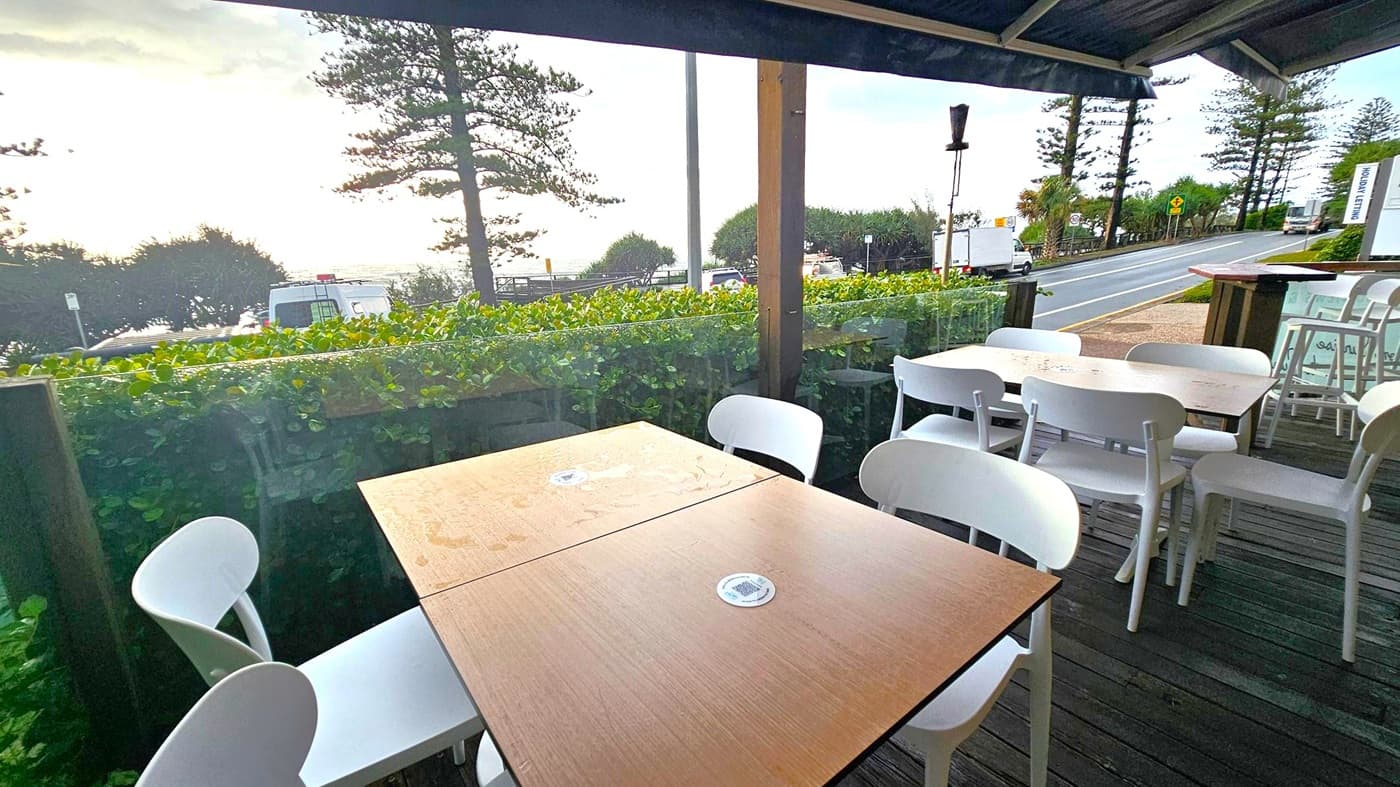 Outdoor seating area with wooden tables and white chairs on a wooden deck. In the background, a road runs parallel to the seating area, with a few vehicles visible. Beyond the road, there are green bushes and tall trees, with the ocean visible in the distance. The sky is overcast, and the area is covered by a roof.