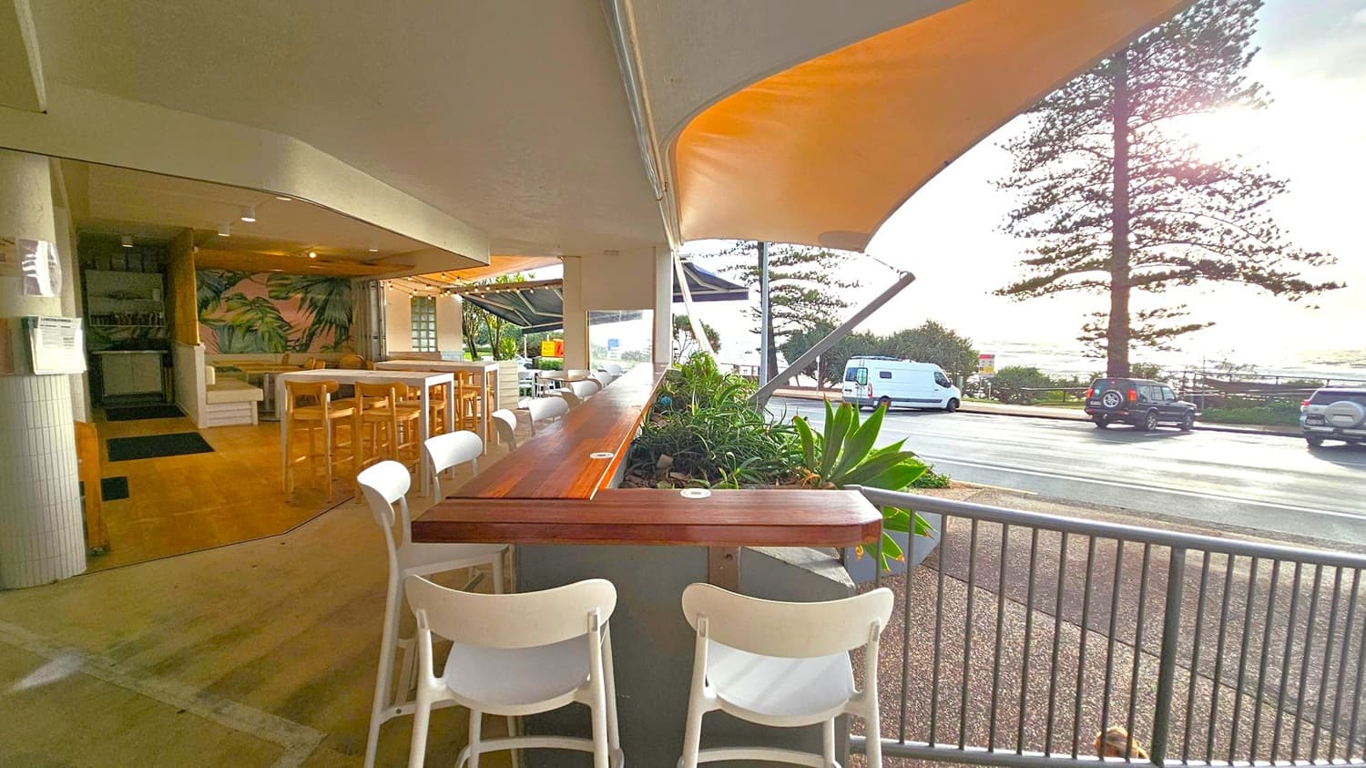 An outdoor café with wooden tables and white chairs on the left, featuring a tropical-themed wall. The seating area is partially covered by a canopy. In the foreground, a wooden counter with plants separates the café from the street. On the right, a road with parked vehicles and a large tree is visible. Beyond the road, the ocean can be seen under a bright sky.