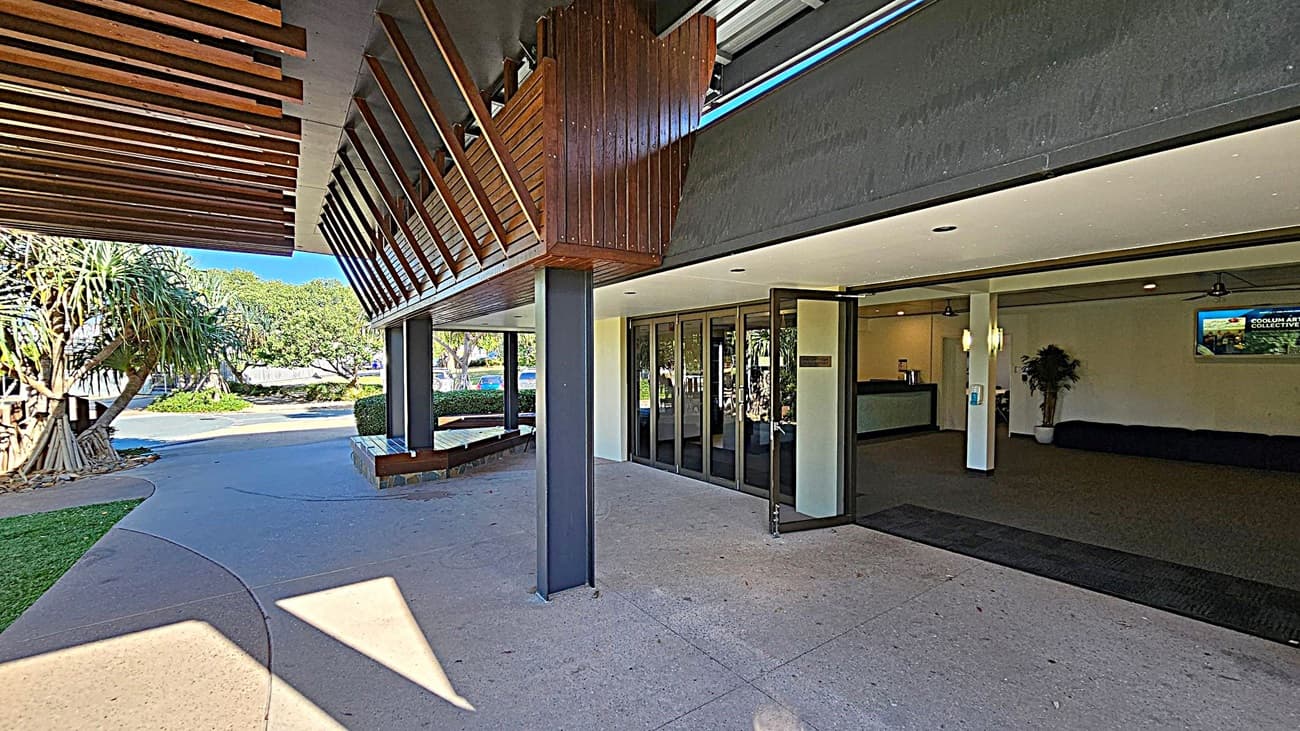 Open double doors lead into a spacious lobby with brown carpet. In the centre, a digital screen is mounted on the wall above a row of black seating. Potted plants are placed on either side. To the right, a hallway with a blue chair and more plants is visible. On the left, a reception desk is partially seen. The ceiling has recessed lighting and wooden panels. A notice board is on the right wall near the entrance.