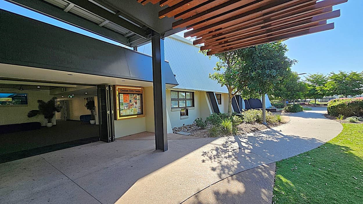 A building entrance on the left with a sign reading "Coolum Civic Centre" and a display board. The interior is dimly lit with potted plants visible. A pathway curves from the entrance to the right, bordered by grass and landscaped bushes. Trees provide shade along the path. The ceiling features wooden slats, and the ground is a mix of concrete and grass.