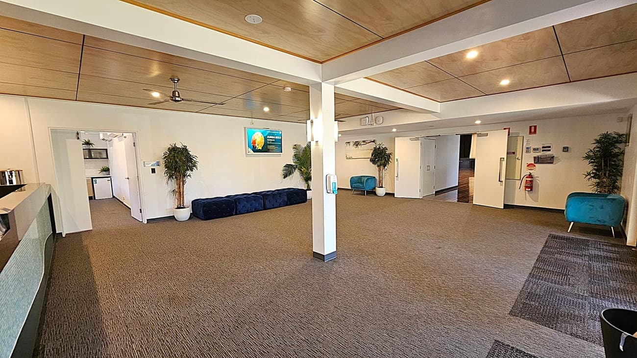 A spacious room with a brown carpeted floor and a wooden panelled ceiling. On the left, there's a reception counter and an open doorway leading to another room with shelves and plants. In the centre, a white column with a hand sanitiser dispenser. The right side features a blue chair, a fire extinguisher, and a potted plant. A digital screen is mounted on the wall, and there are several recessed lights in the ceiling.