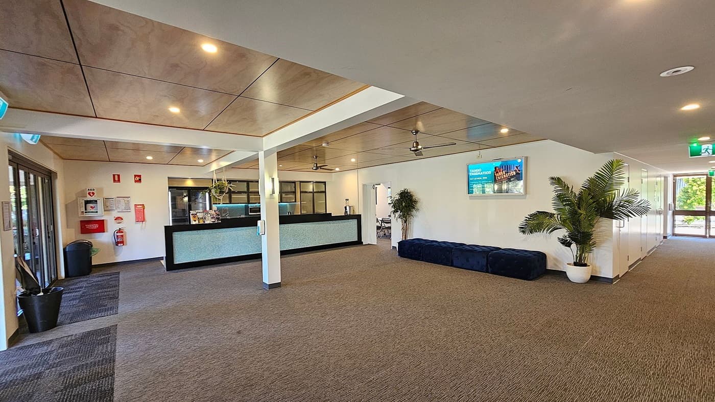 A spacious lobby area with a reception desk on the left, featuring a black counter and a hanging plant above. Behind the desk are windows and a wall with emergency equipment and notices. On the right, a long blue bench sits against the wall, with a large potted plant nearby. A digital screen is mounted above the bench. The floor is carpeted in a mix of grey and brown tones, and the ceiling has recessed lighting and ceiling fans. A glass door is visible at the far end.