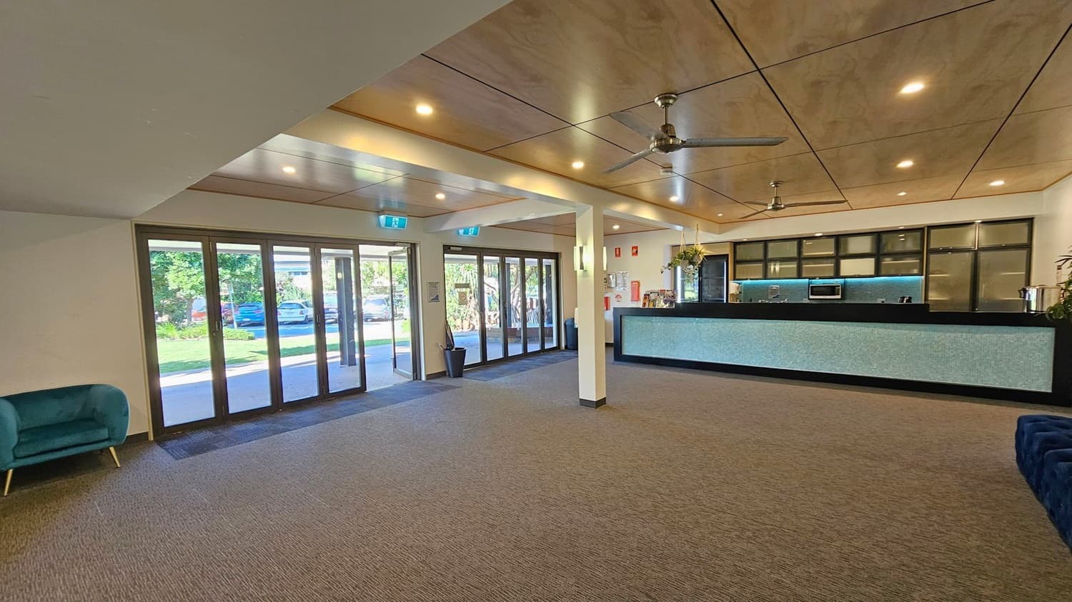 A spacious interior with a carpeted floor and a wooden panelled ceiling featuring recessed lighting and ceiling fans. On the left, there are large glass doors leading outside, with a view of parked cars and greenery. On the right, a long reception desk with a light blue front and black countertop is visible, along with some potted plants. A blue armchair is partially visible in the foreground on the left.