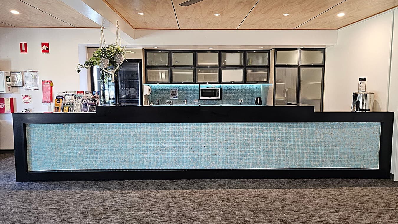 A reception area with a long counter featuring a light blue mosaic front. Behind the counter, there are cabinets with frosted glass doors, a microwave, and a sink. A hanging plant is suspended from the ceiling. On the left, there are brochures and safety equipment, including a fire extinguisher and AED. The ceiling is wooden with recessed lighting, and the floor is carpeted in a grey tone.
