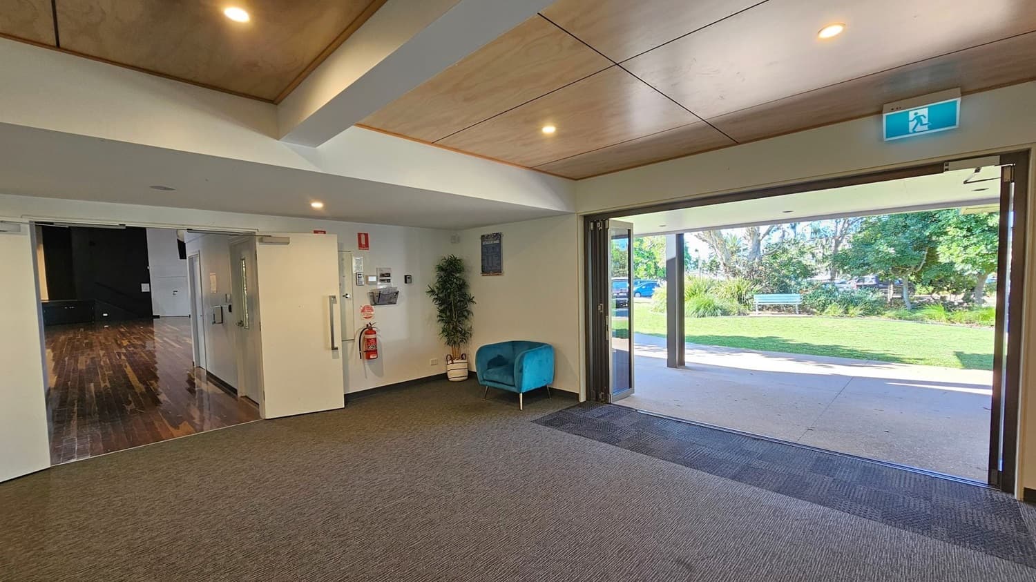 A room with a grey carpet and a wooden ceiling. On the left, there are double doors leading to a room with a wooden floor. A fire extinguisher and a potted plant are near the doors. A blue chair is against the wall. On the right, large glass doors open to a grassy outdoor area with trees and a bench. An exit sign is above the doors.