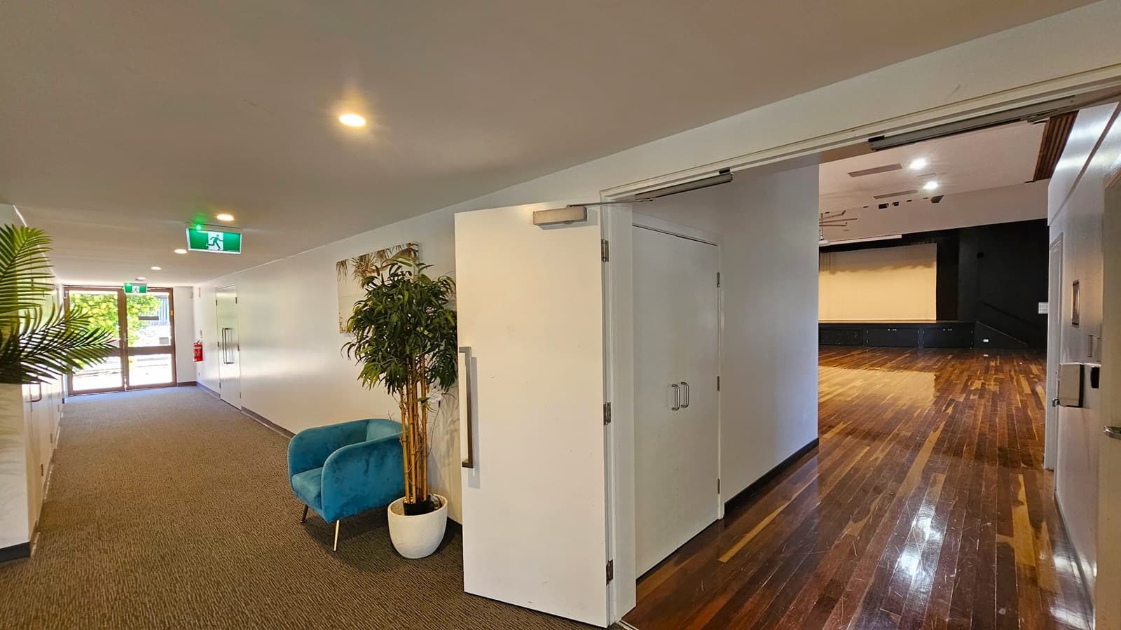 A hallway with a grey carpeted floor and white walls. On the left, there's a green exit sign above a door with glass panels. A potted plant and a blue chair are placed along the wall. On the right, a white door is open, leading to a room with a polished wooden floor and a large screen on the far wall. The ceiling has recessed lighting.