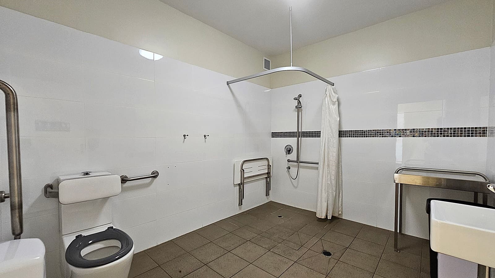 A bathroom with white tiled walls and a brown tiled floor. On the left, there is a toilet with a grey seat and metal grab bars. In the centre, a shower area with a handheld showerhead, a foldable seat, and a white curtain. On the right, a metal-framed sink with a white basin. The ceiling is light green, and there is a small vent above the shower area.