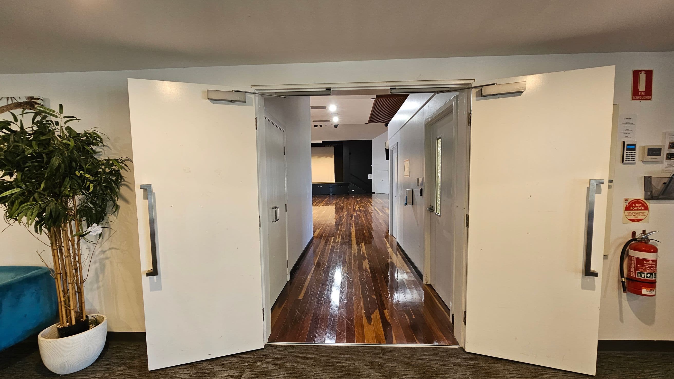 A room with a carpeted floor and a wooden ceiling. On the left, there are double doors leading to a room with a wooden floor. A fire extinguisher and a potted plant are near the doors. A blue chair is against the wall. On the right, large glass doors open to a grassy outdoor area with trees and a bench. An exit sign is above the doors.