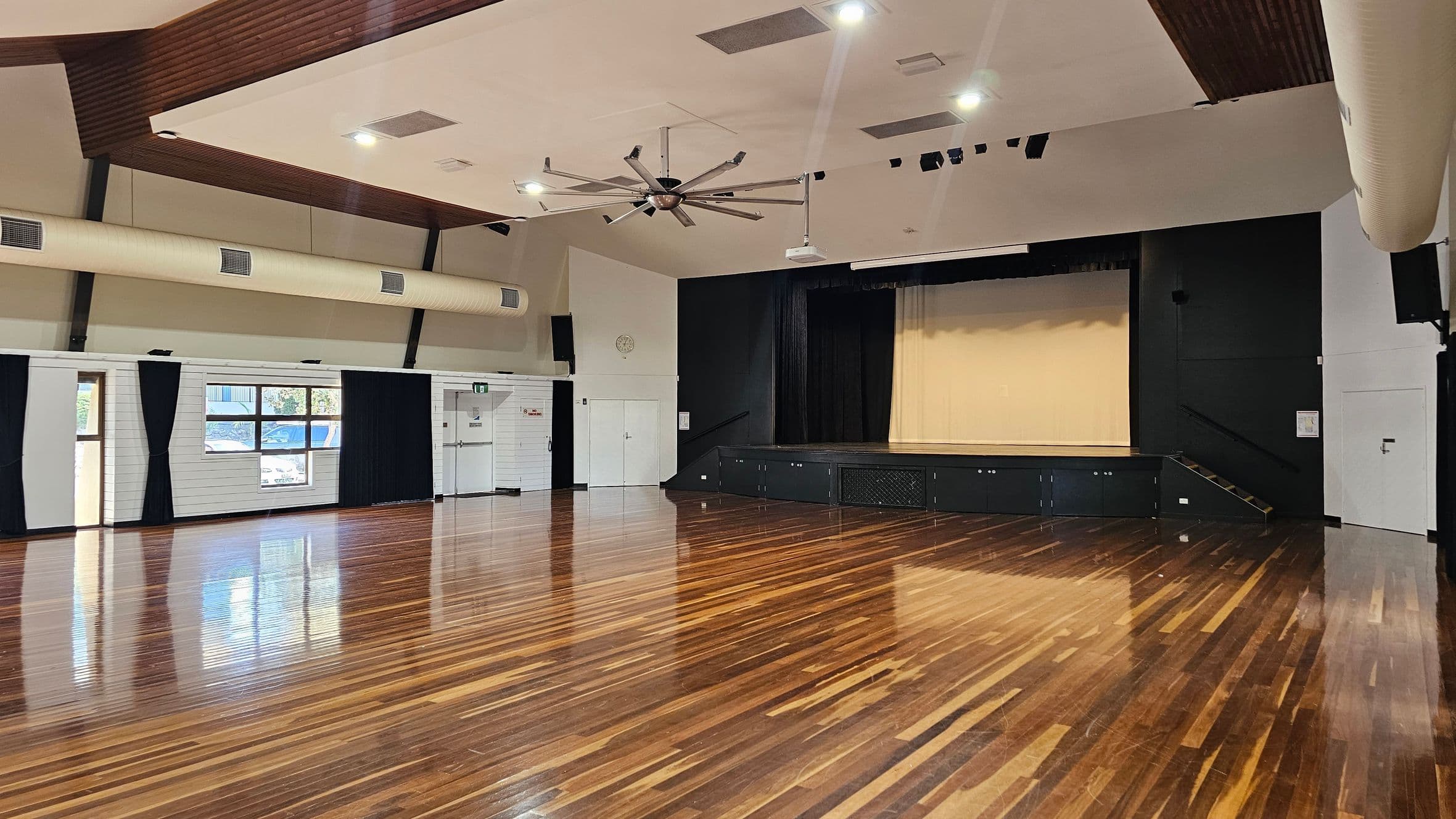 A spacious hall with polished wooden floors and a high ceiling. On the right, there's a stage with a black backdrop and steps leading up to it. The left side features large windows with black curtains, allowing natural light in. The ceiling has a large industrial fan and several recessed lights. The walls are light-coloured with some ventilation ducts visible.