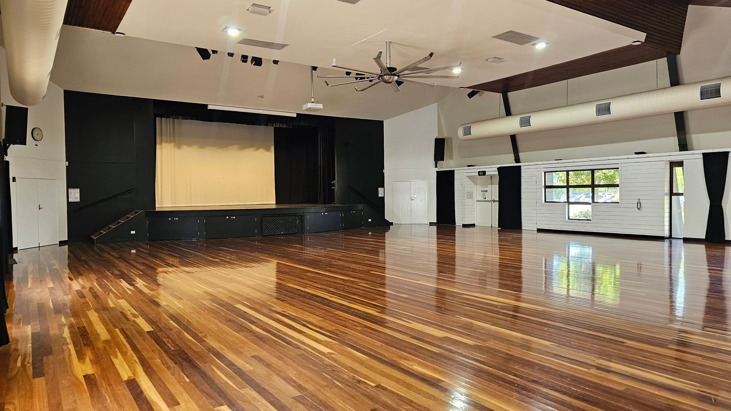 A spacious hall with polished wooden floors and a high ceiling. On the left, there's a stage with a beige curtain and black backdrop. A large ceiling fan is centrally located. The right side features white walls with large windows and black curtains, allowing natural light in. There are air conditioning ducts along the ceiling and a clock on the left wall. The room is empty, highlighting its open space.