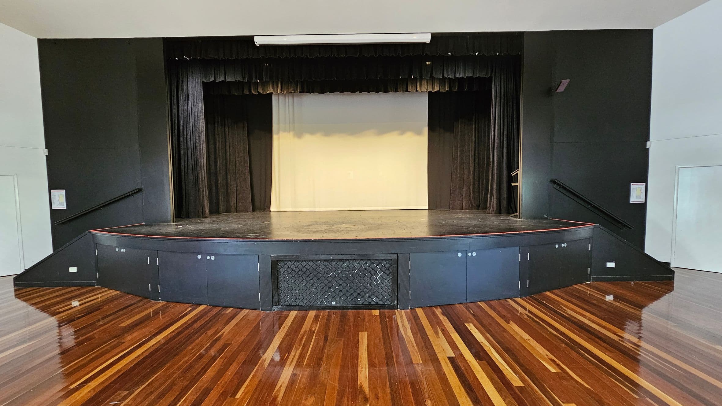 A wooden-floored auditorium with a stage in the centre. The stage has a black backdrop with curtains on either side. The front of the stage has a black panel with a mesh section in the middle. The walls are dark, and there are handrails on both sides leading up to the stage. The ceiling is light-coloured with a white fixture above the stage.