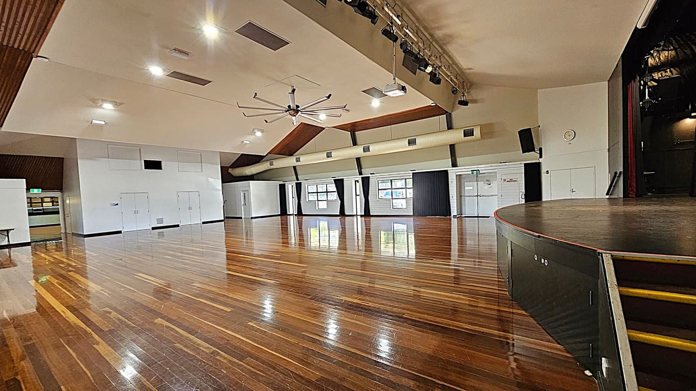 A spacious hall with polished wooden floors and a high ceiling. On the right, there's a stage with a curved edge and steps leading up. The ceiling features a large fan and several lights. The left side has white walls with doors and a small table. The back wall has windows with black curtains and a clock. A ventilation duct runs along the upper part of the wall.