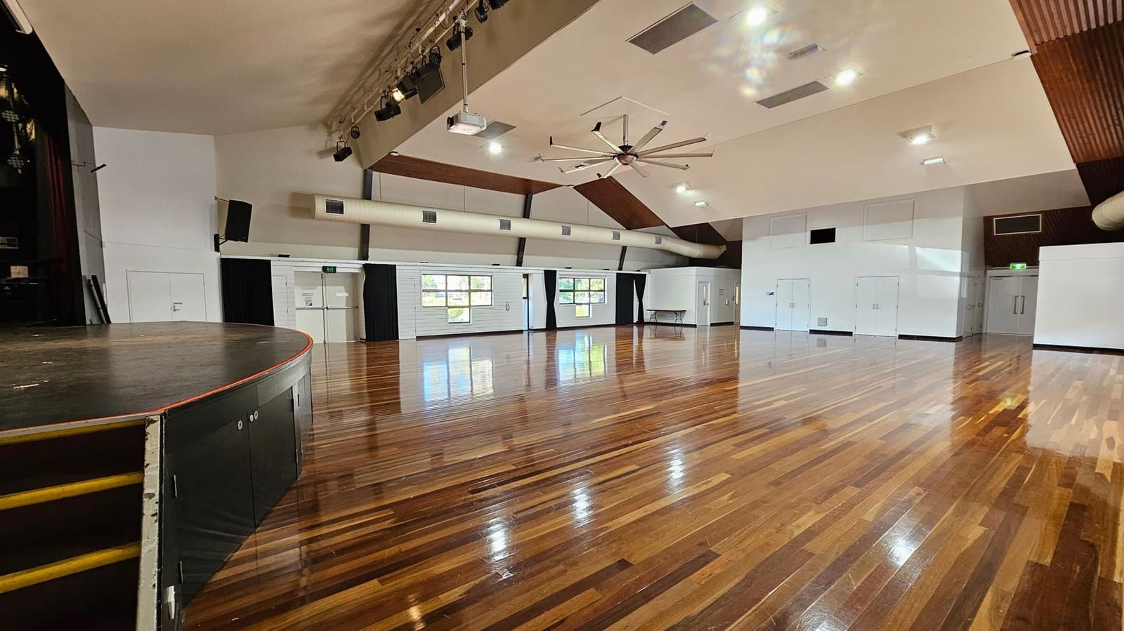 A spacious hall with polished wooden floors and a high ceiling. On the left, there's a stage with steps leading up to it. The ceiling features a large fan and several lights. The back wall has windows with curtains, and there are multiple doors along the walls. A table is positioned against the right wall. The room is well-lit, with reflections visible on the floor.